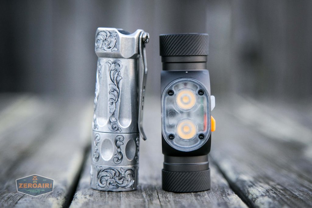 Two small flashlights stand upright on a wooden surface. The left flashlight is silver with decorative engravings and a clip, while the right flashlight is black with two visible LED lights and an orange button.