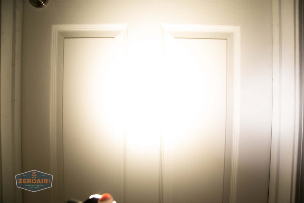 A bright flashlight beam shines directly onto a closed white door, creating a large, intense spot of light in the center. A small ZeroAir logo is visible in the lower left corner of the image.