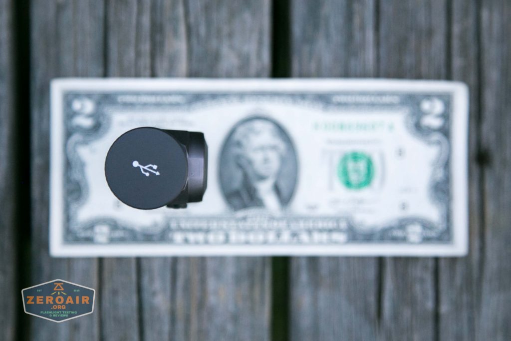 A USB connector facing upward is placed on top of a U.S. two-dollar bill laid flat on a wooden surface. The two-dollar bill is mostly visible, with the connector partially blocking Thomas Jefferson’s portrait.