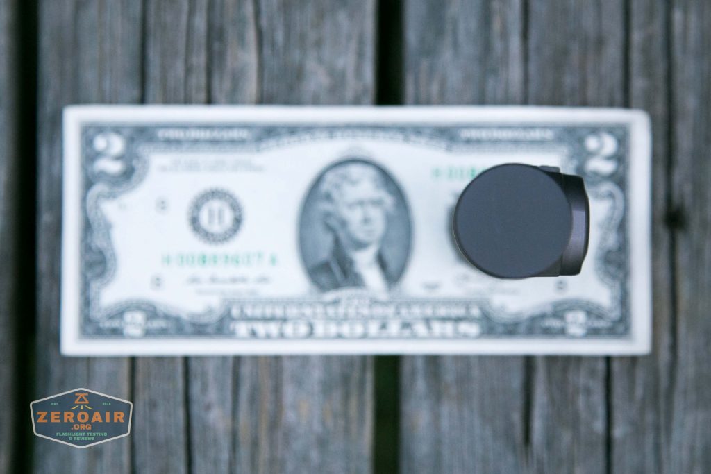 A two-dollar bill lies flat on a wooden surface with a round, black object placed vertically on top of its right side, partially covering the bill. A ZeroAir logo is in the bottom left corner.