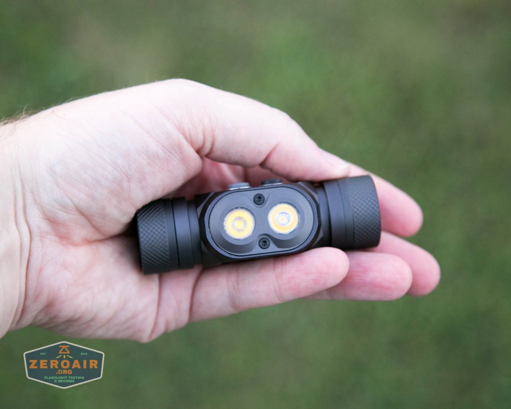 A hand holds a black flashlight with two round LED bulbs visible at the front. The flashlight has a textured grip on both ends. The background is out of focus grass. A ZeroAir logo is in the lower left corner.