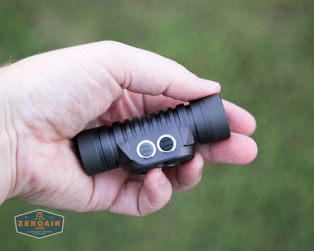 A hand holding a small black flashlight with two silver buttons on top. The background is out of focus green grass. A logo in the bottom left corner reads ZEROAIR.