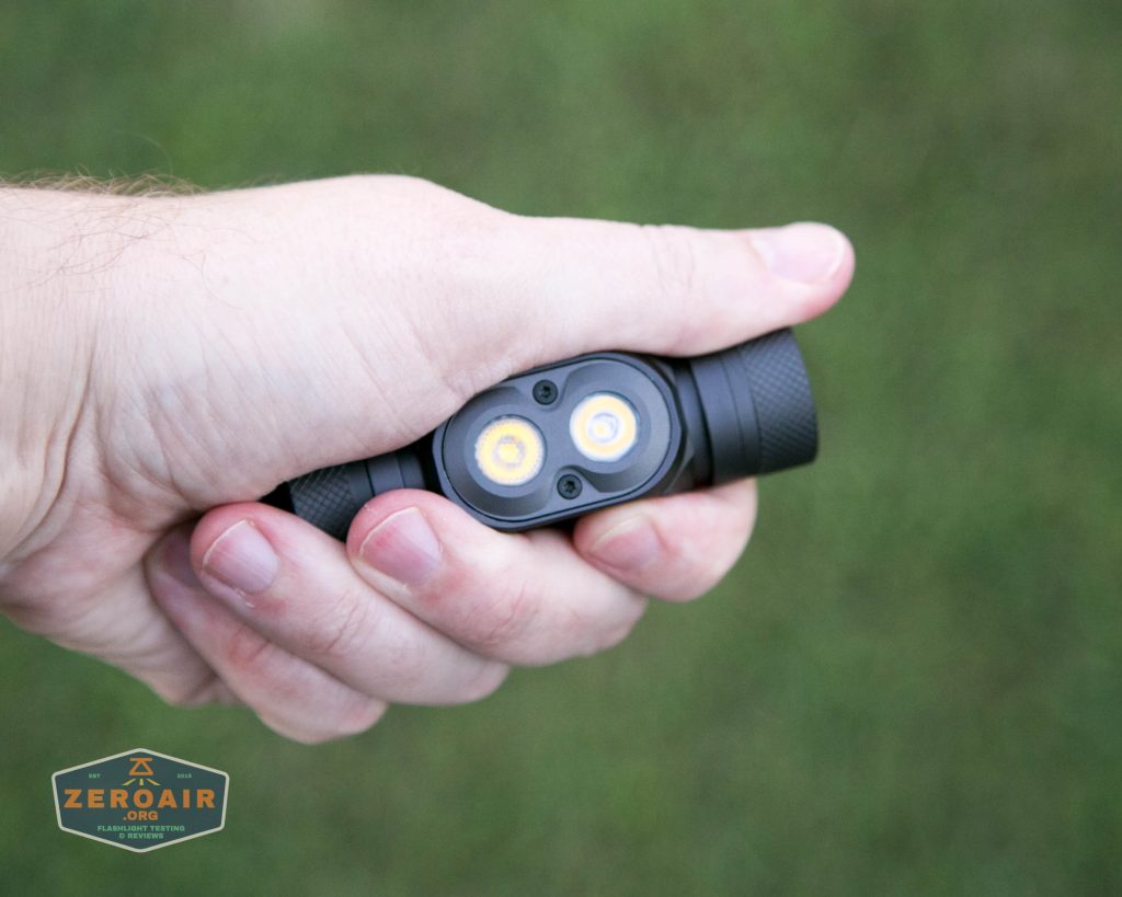 A hand holding a small, black flashlight with two circular LED bulbs. The background is green and out of focus. A logo reading ZeroAir is visible in the bottom left corner.