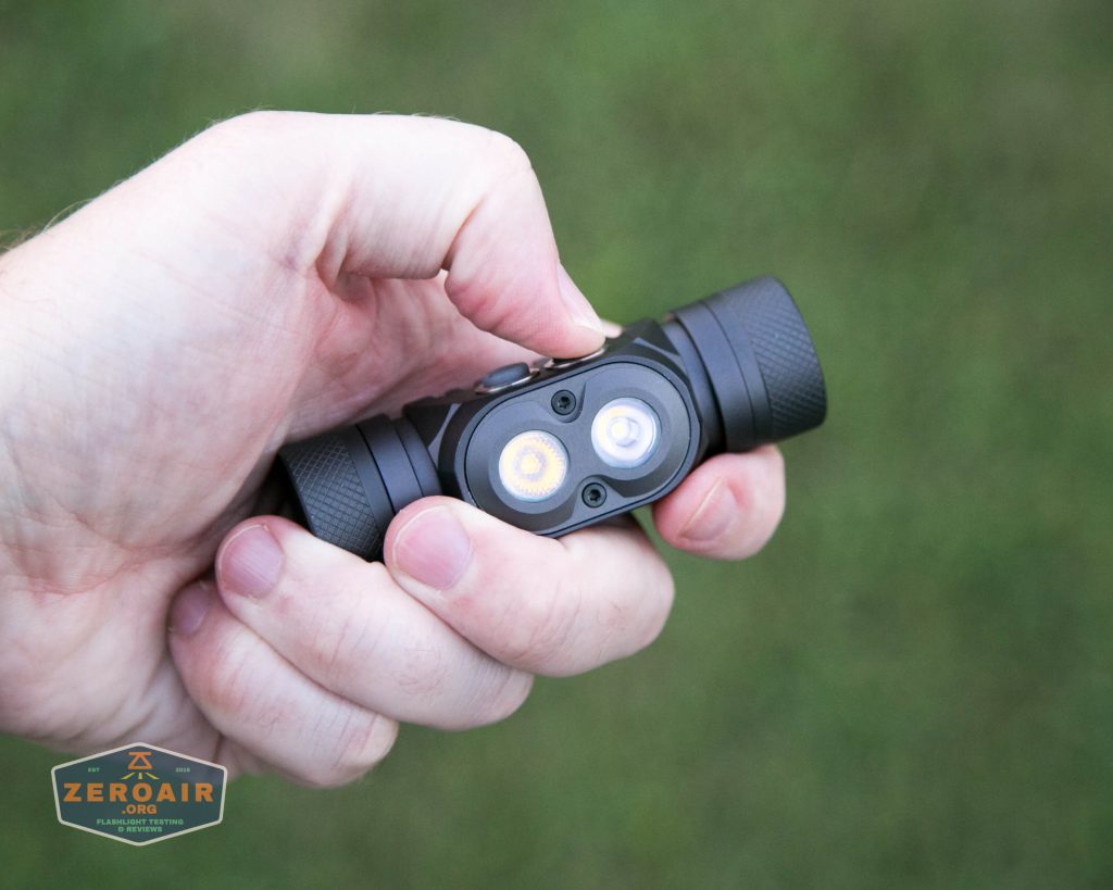 A hand holds a small, black flashlight with two LED bulbs, one yellow and one white, against a blurred green background. A small ZeroAir logo appears in the lower left corner.