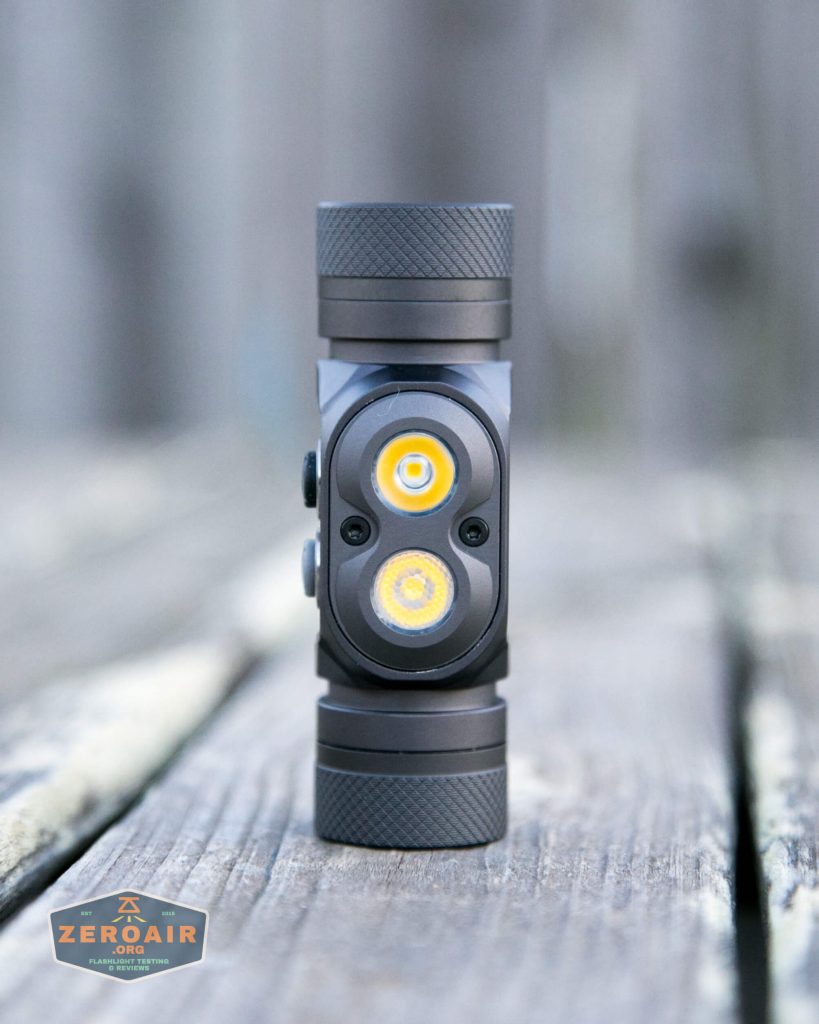 A small black flashlight with two circular LED lights and one smaller circle in between, standing upright on a wooden surface. The background is blurred, and a ZeroAir logo is visible in the bottom left corner.