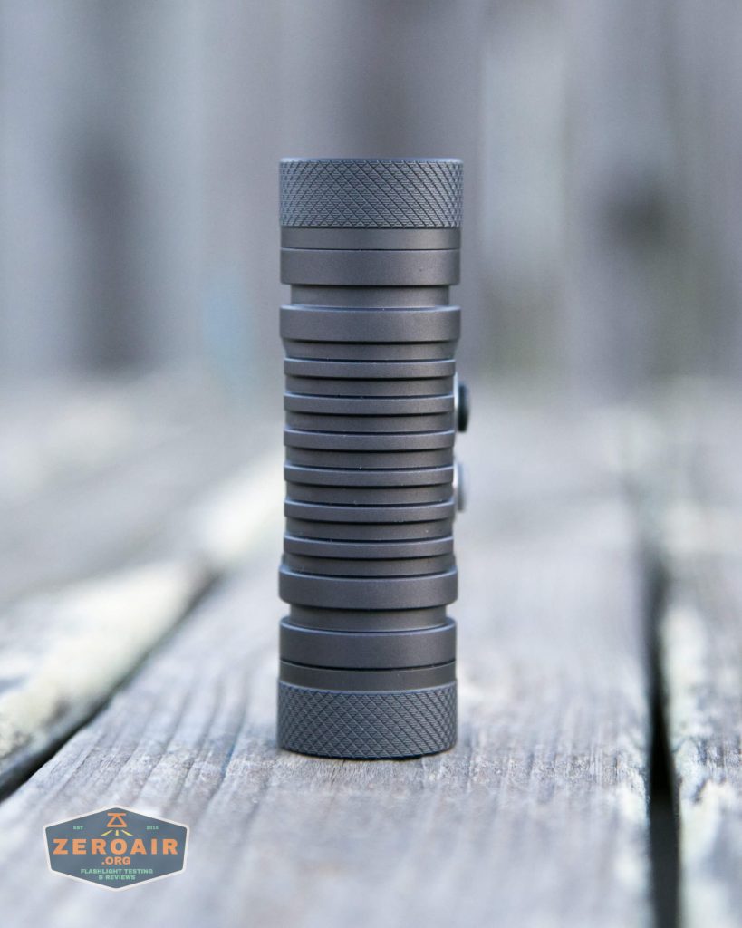 A cylindrical, dark gray flashlight with a ribbed body stands upright on a weathered wooden surface. A small ZeroAir logo appears in the lower left corner of the image.