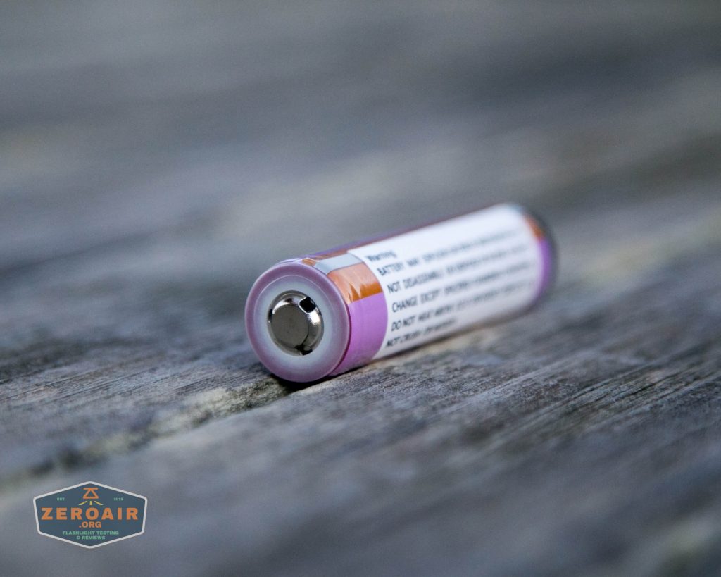 A close-up of a single cylindrical battery lying horizontally on a weathered wooden surface, with a partially visible label and the ZEROAIR logo in the bottom left corner.
