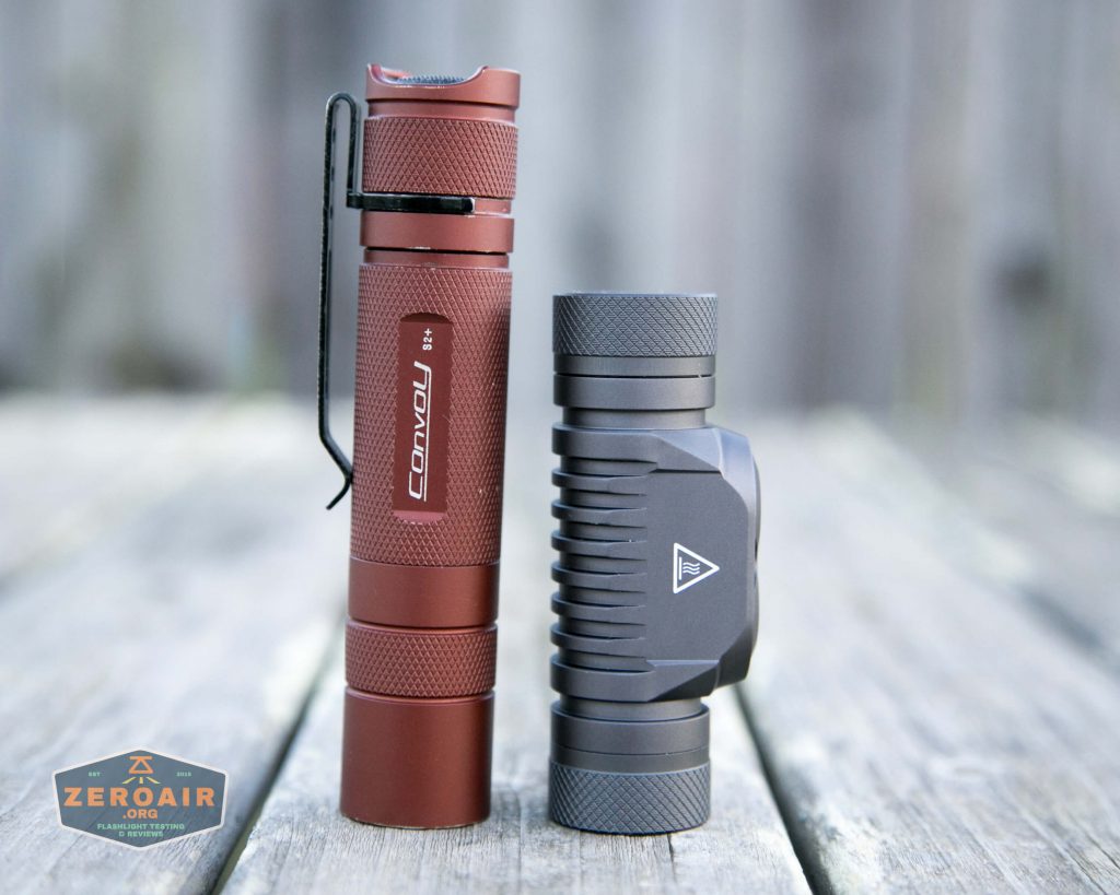 Two compact flashlights stand upright on a weathered wooden surface. The flashlight on the left is red with a black clip and Convoy written on the side. The flashlight on the right is gray with a textured grip.