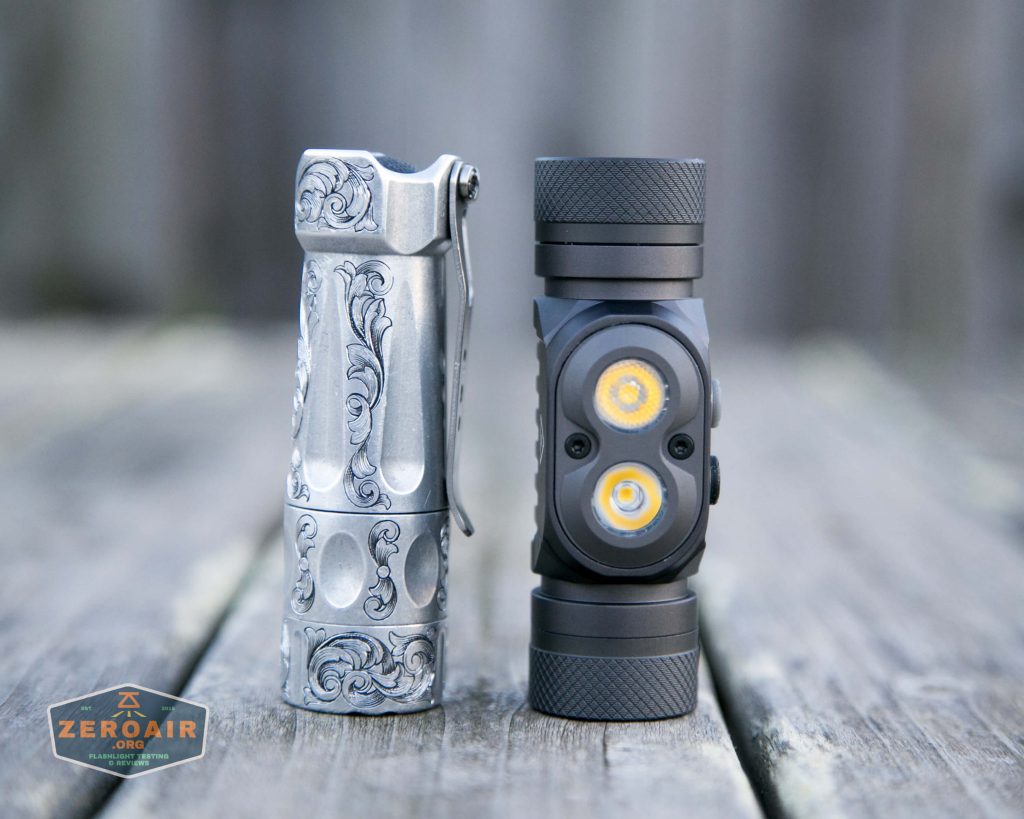 Two compact flashlights stand upright on a weathered wooden surface. The left flashlight has ornate engravings and a pocket clip, while the right one is matte black with two exposed LED lights. A small ZeroAir logo is in the lower left corner.