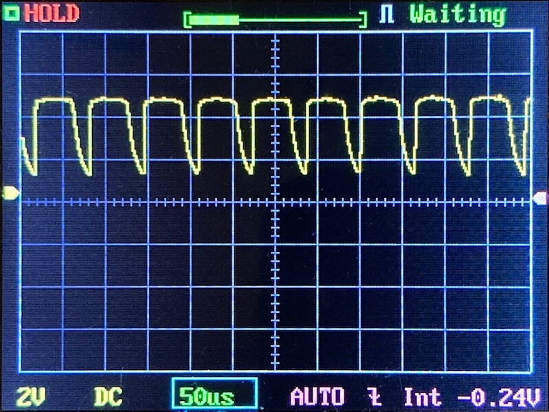 An oscilloscope screen displays a repeating yellow waveform with sharp peaks and dips. The settings show 2V per division vertically and 50 microseconds per division horizontally. Status indicators and grid lines are visible.