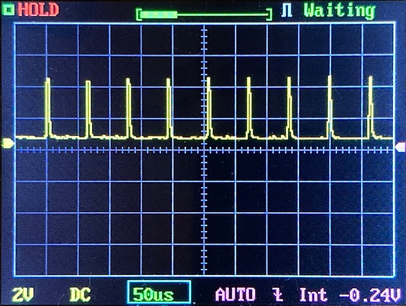 A digital oscilloscope screen displays a repeating series of sharp, narrow voltage pulses. The settings show 2V per division vertically and 50 microseconds per division horizontally. The screen shows HOLD and Waiting status.