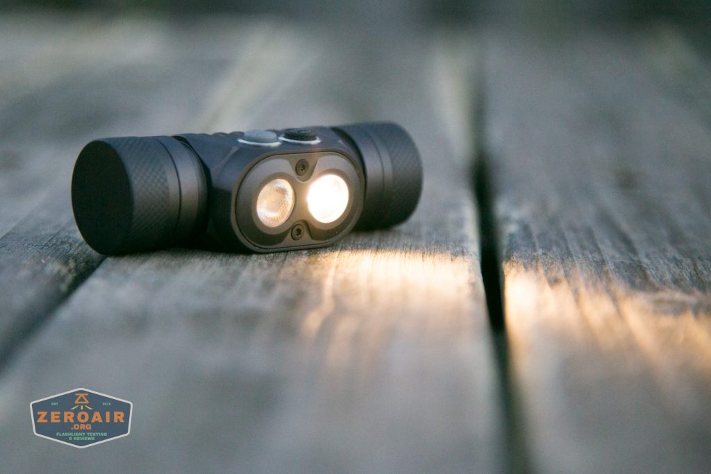 A compact black flashlight with two illuminated bulbs rests on a weathered wooden surface. A ZeroAir logo is visible in the bottom left corner of the image.