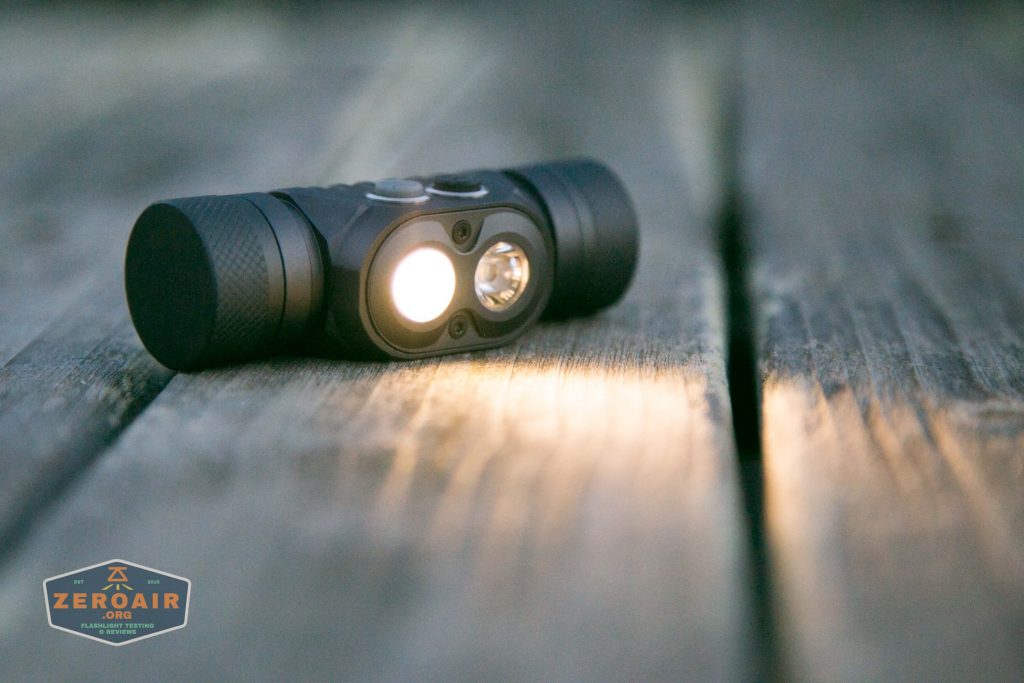 A black flashlight with two lenses, one illuminated, rests on a wooden surface. Light from the flashlight shines onto the wood. The ZeroAir logo appears in the bottom left corner.