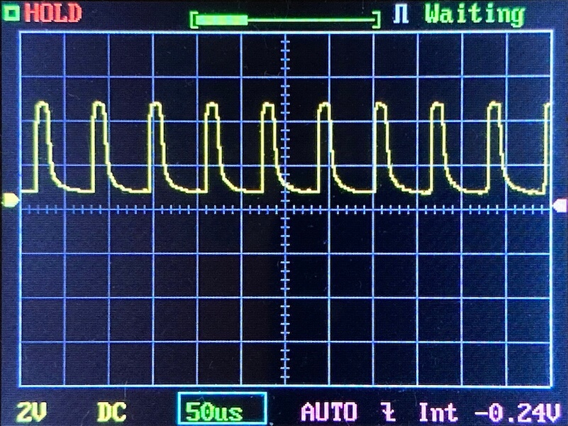 Oscilloscope screen displaying a repeating yellow waveform with sharp peaks and gradual declines. The settings at the bottom read 2V, DC, 50µs. The top right indicates the status as “Waiting.”