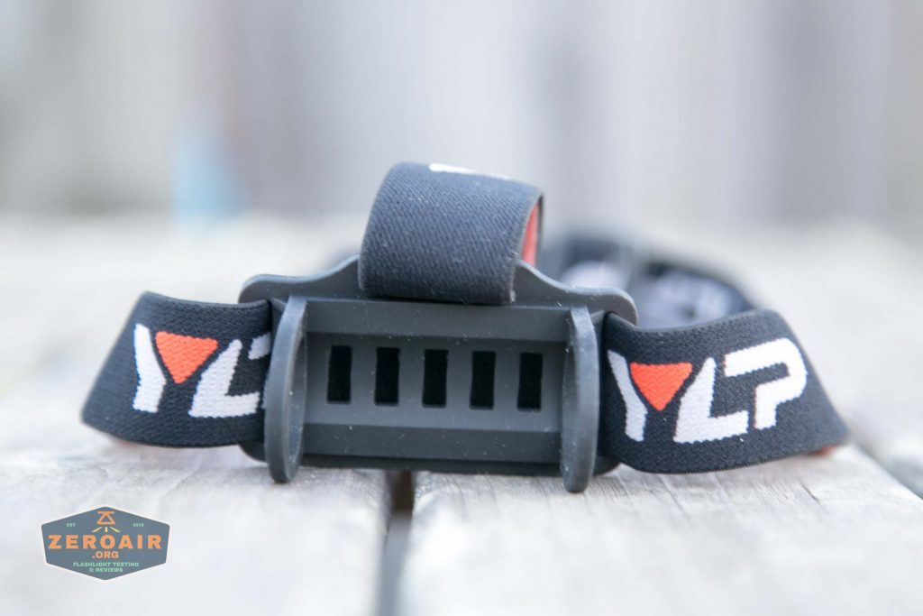 A close-up view of a black headlamp strap with bold white letters YLP and an orange triangle logo, attached to a plastic bracket. The background is blurred.