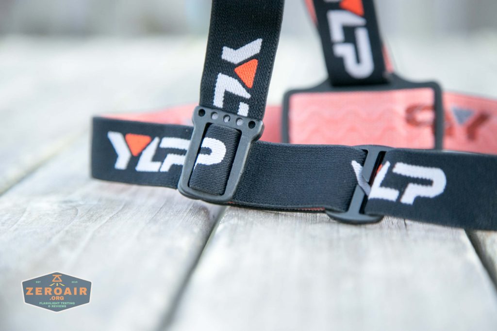 A close-up of a black and orange adjustable strap with “YLP” branding, laying on a wooden surface. The focus is on the plastic buckle and the logo. A ZeroAir watermark is in the lower left corner.