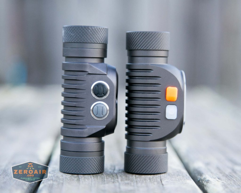 Two cylindrical, dark gray devices with textured ends are standing upright side by side on a wooden surface. Each has buttons on the side; one device has two silver buttons, the other has an orange and a gray button.