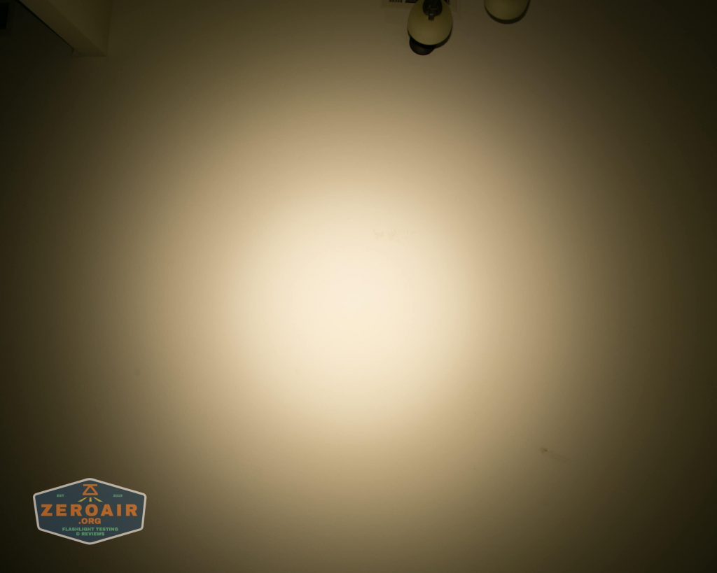 A blank wall is illuminated by a circular beam of light. Two small ceiling light fixtures are partially visible at the top right. A sticker with the text “ZEROAIR” is in the bottom left corner.