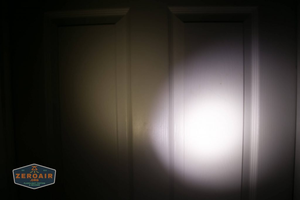A white door is illuminated by a bright circular beam of light in a dark environment. The edges of the door and surrounding area are partially visible, with a logo in the lower left corner.