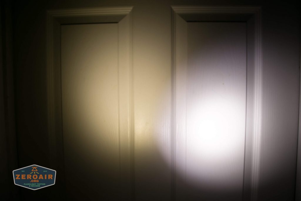 A white door with two panels is illuminated by the beam of a flashlight, creating a circular patch of light on the right side. A small logo in the lower left corner reads ZEROAIR.