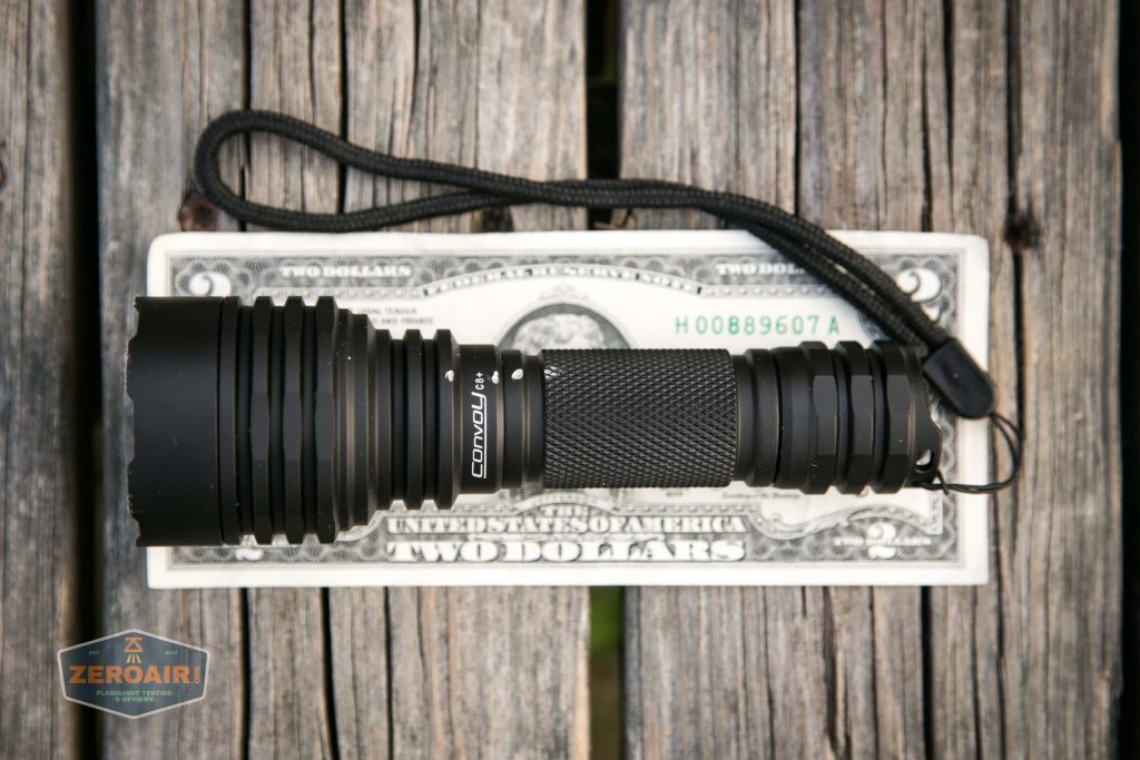 A black Convoy L6 flashlight with a wrist strap is placed on top of a two-dollar bill on a weathered wooden surface. A small ZeroAir logo appears in the bottom left corner.