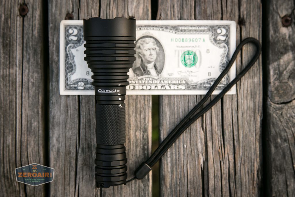 A black Convoy flashlight with a wrist strap is placed on a two-dollar bill, which lies on a weathered wooden surface. A ZeroAir logo appears in the bottom left corner.