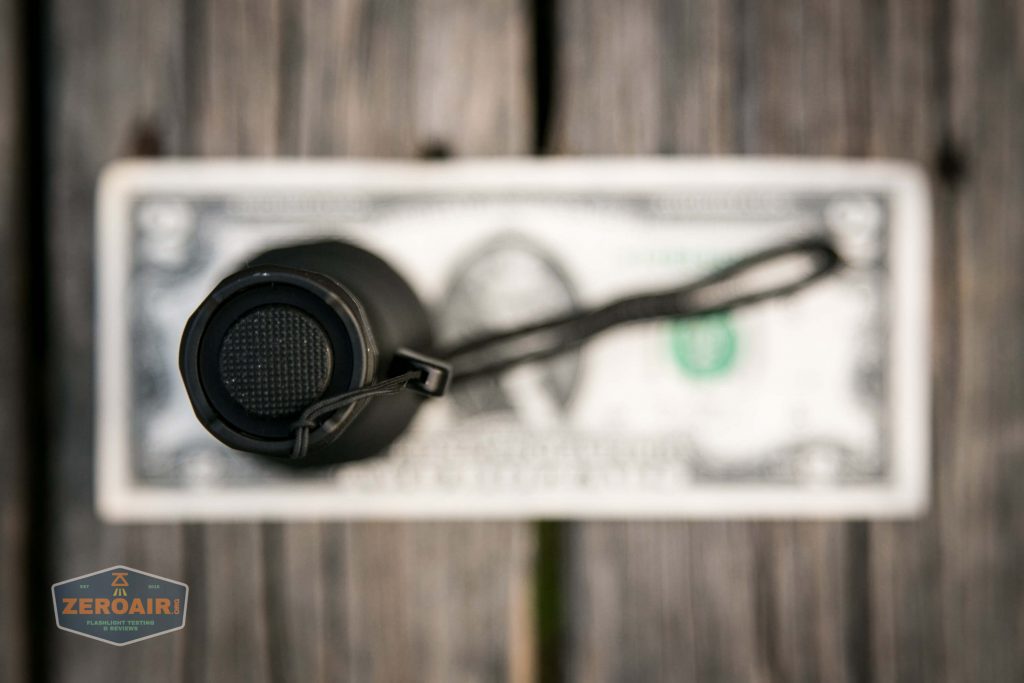 A black cylindrical object with a textured button on top is placed vertically on a US two-dollar bill. A black lanyard is attached, and the bill rests on a wooden surface. The image is slightly blurred.