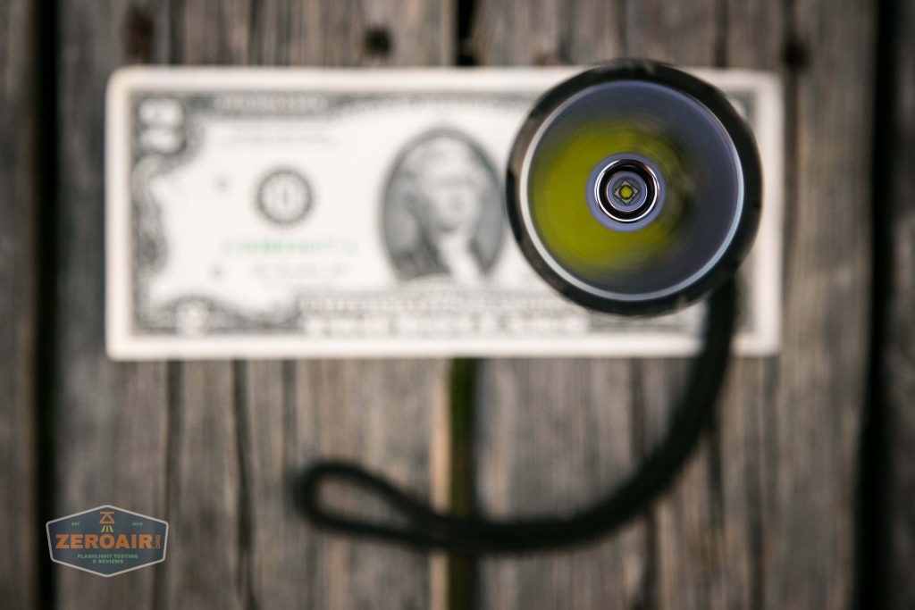 A close-up, top-down view of a flashlight facing up, positioned above a U.S. two-dollar bill placed on a wooden surface. The flashlight’s lanyard is visible, curving to the right. A ZeroAir! logo appears bottom left.