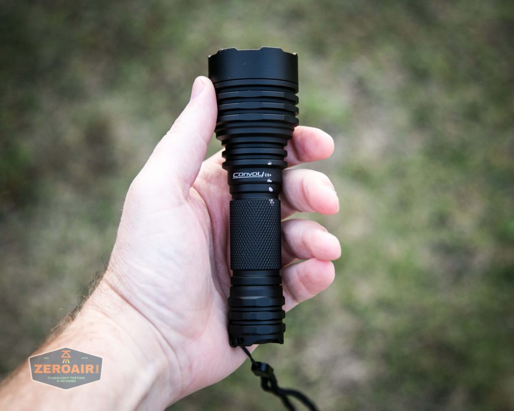 A person holding a black Convoy L21A flashlight outdoors with a grassy background. The flashlight has a ribbed head and a textured grip. The ZeroAir logo is visible in the lower left corner.