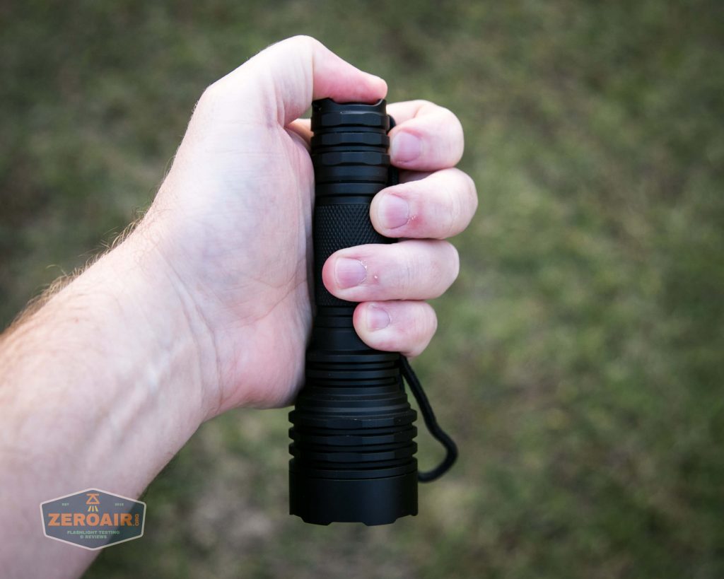 A person holds a black flashlight in their right hand over a blurred grassy background. The flashlight has a textured grip and a lanyard attached. A small ZeroAir logo appears in the lower left corner.
