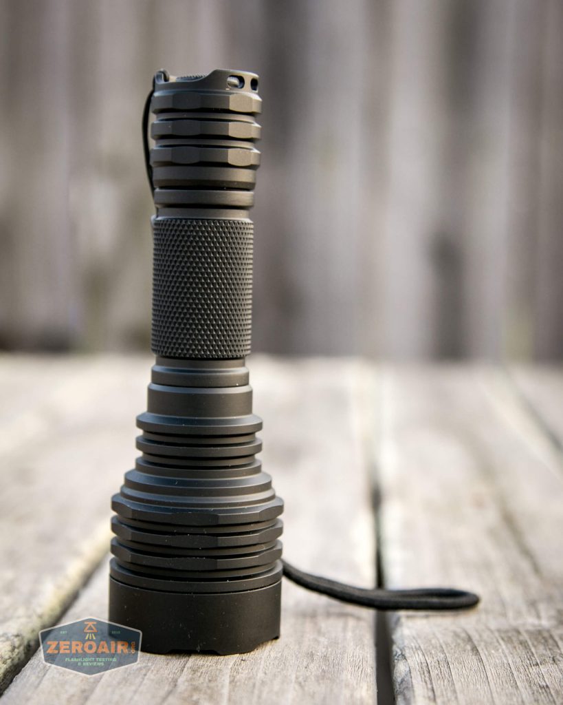 A black, textured flashlight with a wide, ridged head and a wrist strap sits upright on a weathered wooden surface, with a blurred wooden fence in the background.