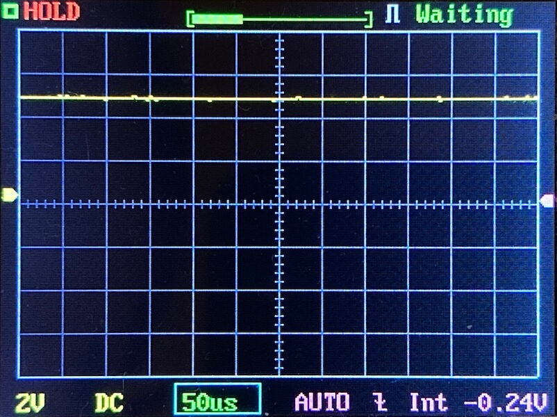 A digital oscilloscope screen displays a flat yellow waveform near the top with grid lines. The settings show 2V per division, 50 microseconds per division, and a DC coupling. Status indicators read “HOLD” and “Waiting.”