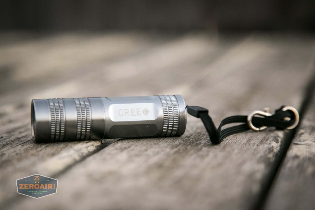 A small silver flashlight with CREE printed on its side rests on a wooden surface. The flashlight has a black wrist strap attached. The ZeroAir logo appears in the bottom left corner.