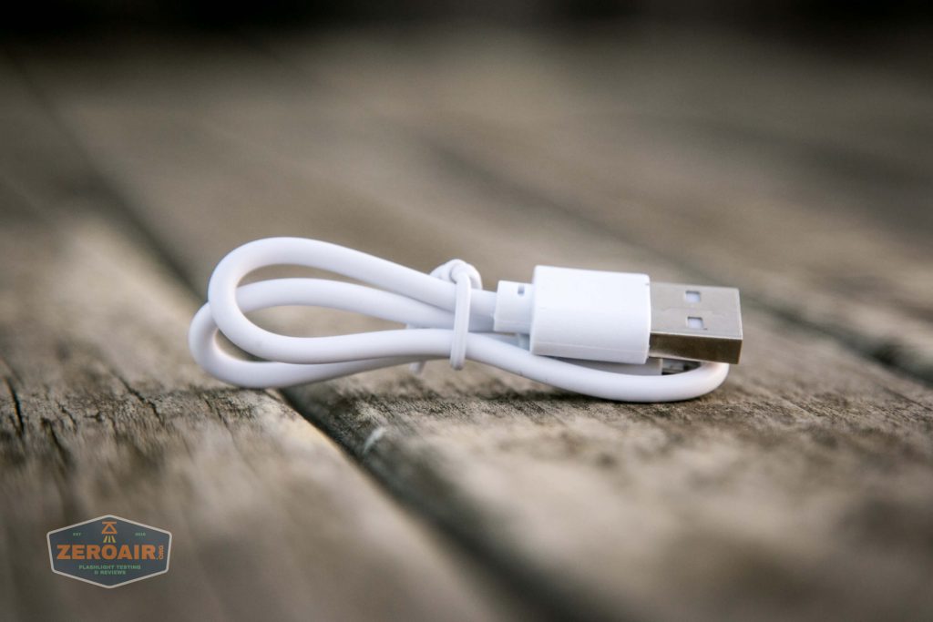 A white USB cable is coiled and secured with a twist tie, resting on a wooden surface. The cable features a standard USB-A connector. A small ZEROAIR logo appears in the lower left corner.
