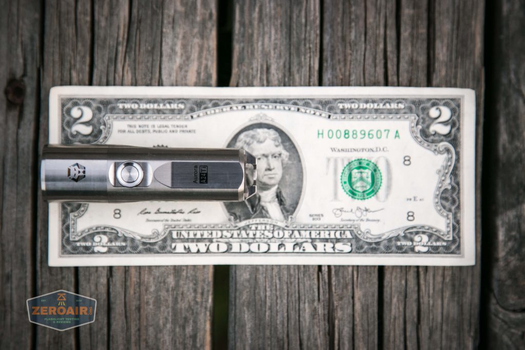 A stainless steel flashlight placed horizontally on top of a U.S. two-dollar bill, covering the left side of the bill. The bill is on a wooden surface, and a ZeroAir watermark is in the lower left corner.