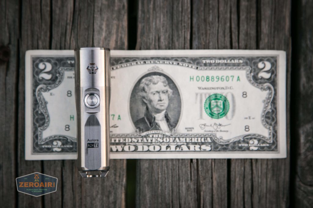 A small silver flashlight is placed vertically on top of a two-dollar bill, which is lying on a weathered wooden surface. A logo reading “ZEROAIR” appears in the lower left corner.