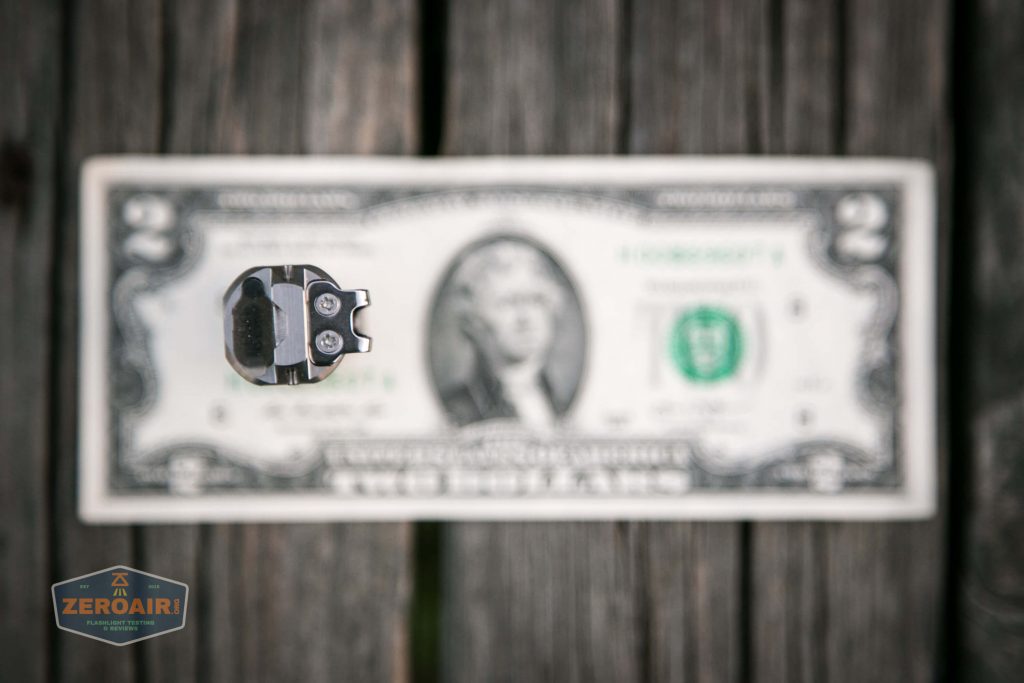 A small metallic object is in sharp focus in the foreground, with a blurred two-dollar bill and a wooden surface in the background. A ZeroAir logo appears in the lower left corner.