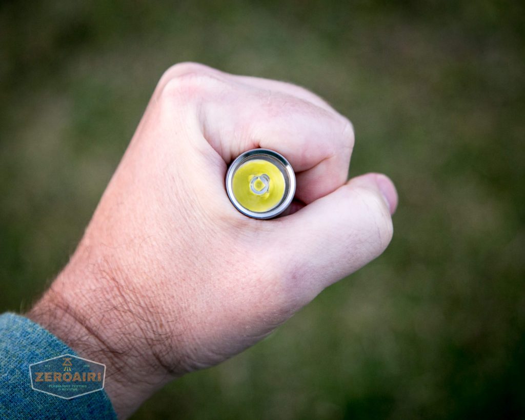 A hand grips a cylindrical object, likely a flashlight, viewed from above with the light facing the camera. The background is out of focus and appears to be grass. A logo that reads Zeroair is visible in the lower left corner.