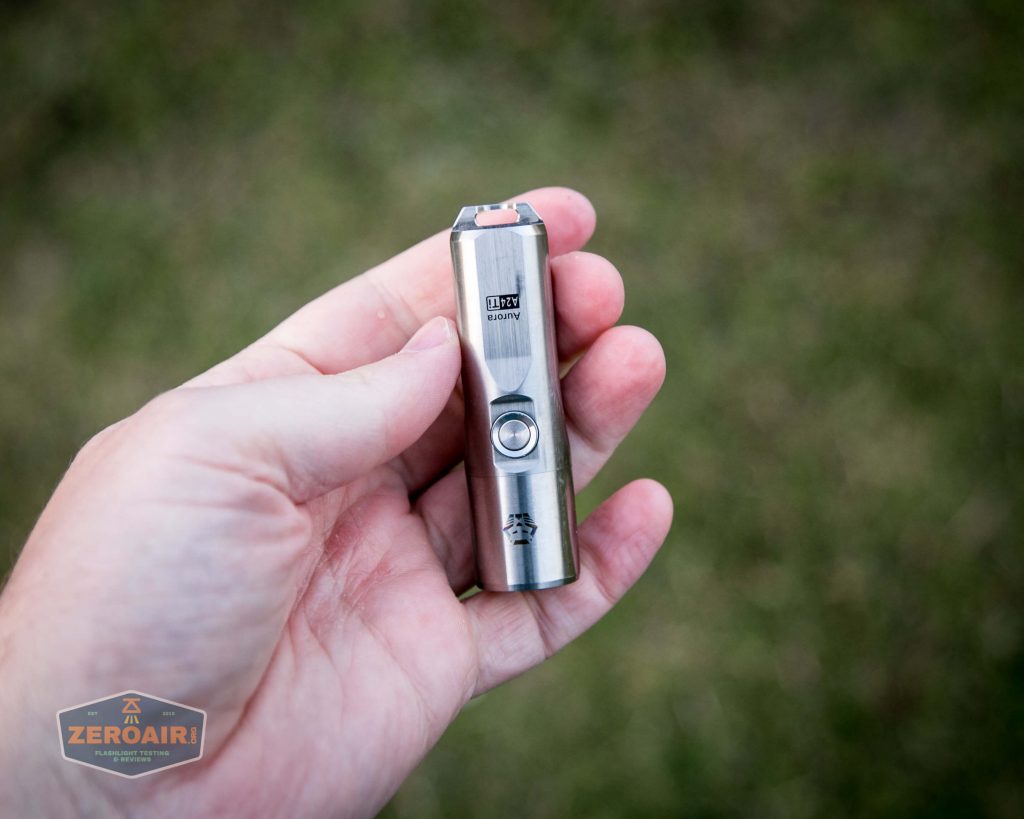 A hand holds a small, stainless steel flashlight with a button and engraving on its body. The background is a blurred grassy area. A ZeroAir watermark is visible in the lower left corner.
