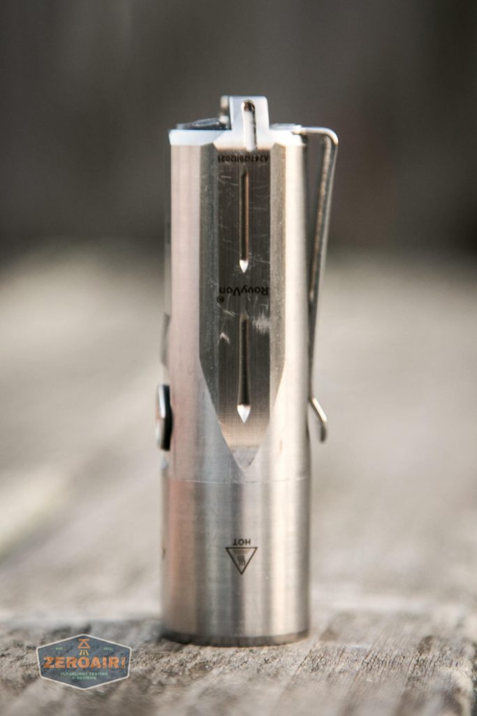 A stainless steel cylindrical flashlight with a pocket clip stands upright on a wooden surface. The device has etched markings and a triangular caution symbol near the base. The background is blurred.