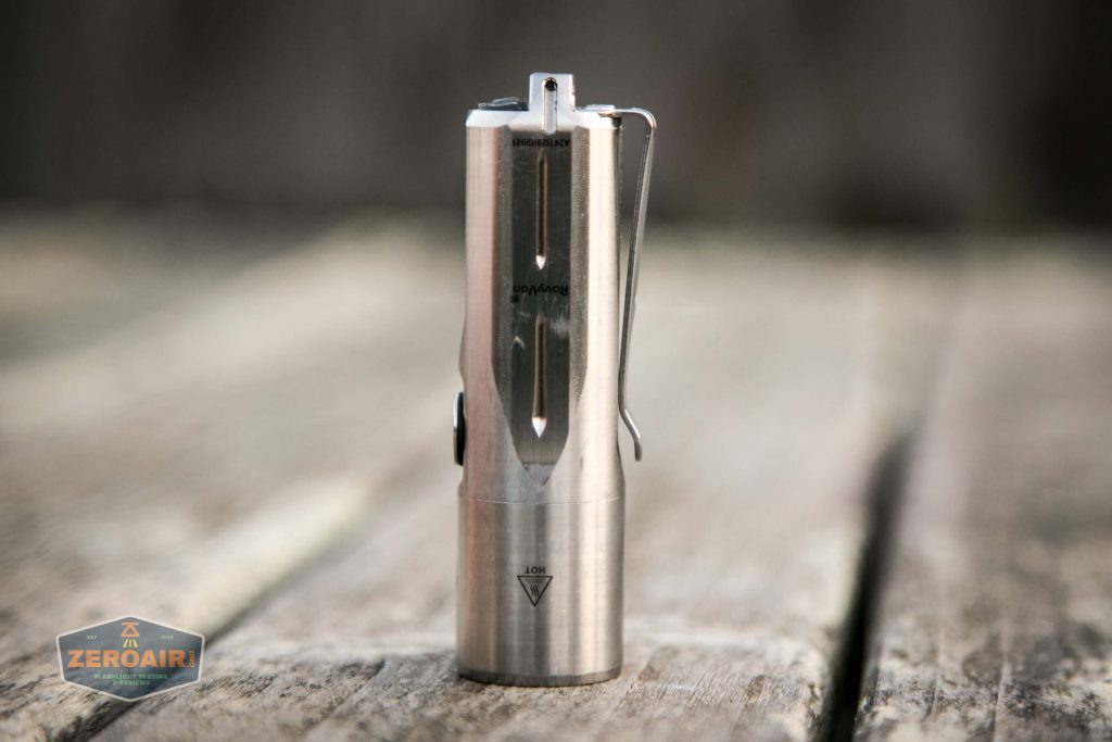 A small stainless steel flashlight standing upright on a wooden surface outdoors, with a clip attached to its side and engraved markings visible on its body.