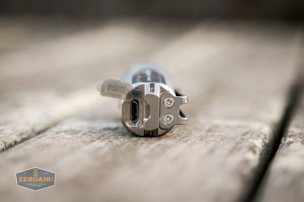 A close-up view of a flashlight lying on a wooden surface, showing the USB charging port with its rubber cover open.