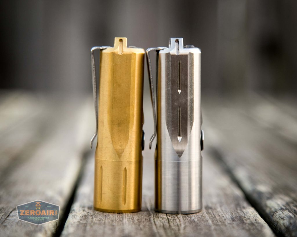 Two small cylindrical flashlights stand upright side by side on a wooden surface. One flashlight is brass-colored and the other is silver. Both have metal clips and arrow markings. The background is blurred.