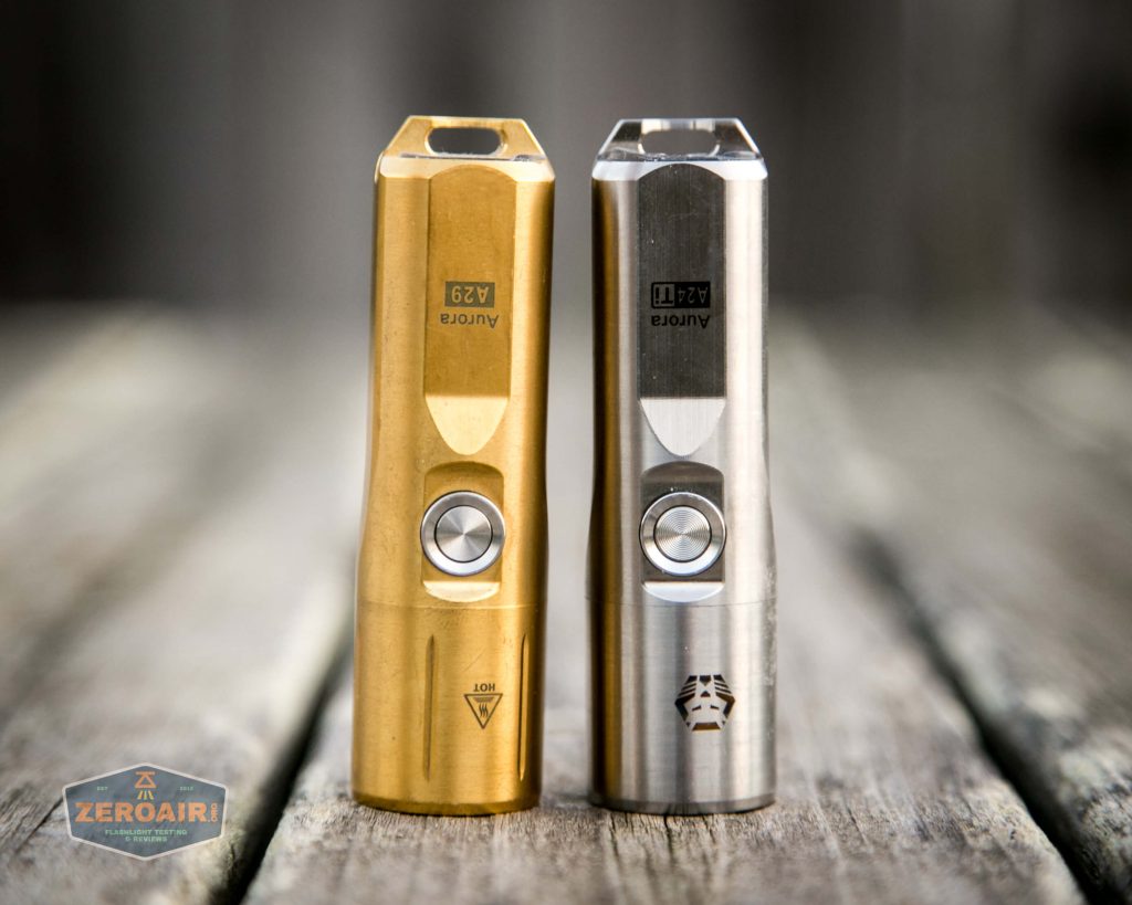 Two metallic, cylindrical flashlights—one gold-colored and one silver—stand upright side by side on a wooden surface. Each has a button on the front; the silver one has a logo near the base. A Zer0Air logo appears in the lower left.