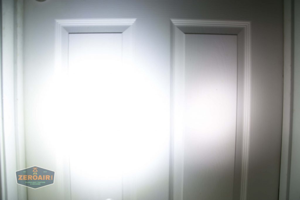 A white door with rectangular panels is illuminated by a bright, circular flashlight beam at the center. A small sticker with the text “ZEROAIR!” is visible in the lower left corner.