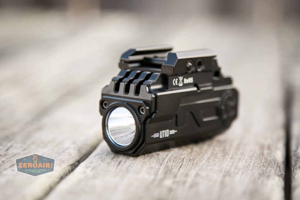 A compact, black tactical flashlight with rails and branding on the side, placed on a weathered wooden surface. The logo ZEROAIR is visible in the bottom left corner.