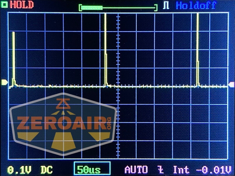An oscilloscope screen displays a signal with two sharp pulses on a grid. Indicators show HOLD, Holdoff, and settings: 0.1V DC, 50μs, AUTO, and -0.01V. A ZEROAIR.org watermark is present.