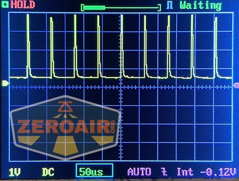 A digital oscilloscope display shows a repeating square wave signal with sharp peaks. The screen includes a grid and the settings: 1V, DC, 50µs. A Zeroair.org logo is visible in the lower left corner.