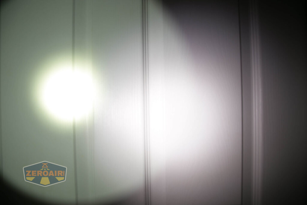 A bright circular beam of light is projected onto a white paneled surface, with the ZeroAir logo partially visible in the lower left corner. The light creates a strong contrast against the background.