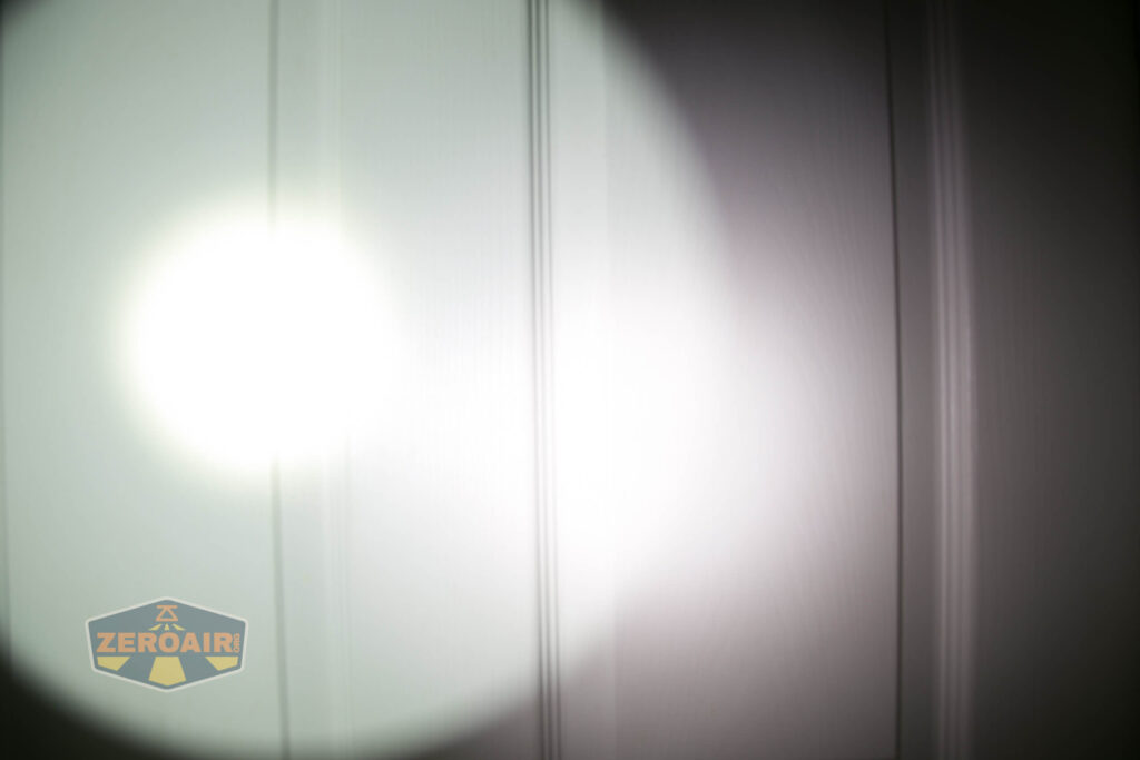 A round beam of flashlight shines on a white paneled door, creating a bright circular spot and softer outer glow. A “ZeroAir” logo appears in the lower left corner of the image.
